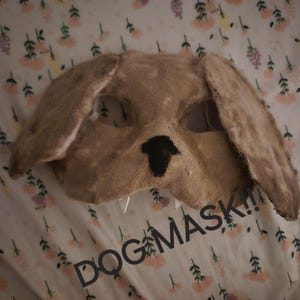 May include: A brown dog mask with a black nose and white teeth. The mask is made of felt and has a distressed look. The text "DOG MASK!!" is written in black letters on the bottom of the mask.
