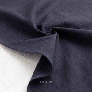 May include: Close-up of a dark navy blue fabric with a textured weave. The fabric is draped and folded, creating shadows and highlighting the material's texture. The word "FABLEISM" is printed in white at the bottom right corner.