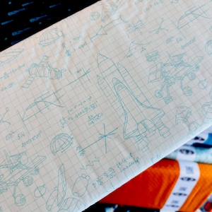 May include: A light blue and white fabric with a space theme. The fabric features illustrations of rockets, satellites, and mathematical equations. The background is a grid pattern. Other fabrics with different patterns are visible in the background.