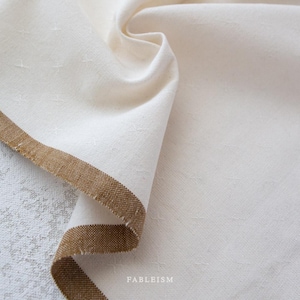 May include: Cream-colored linen fabric with a brown border. The fabric is folded, showing texture and subtle star-like patterns. The word "FABLEISM" is printed in the bottom right corner.