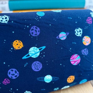 May include: Dark blue fabric with a repeating pattern of colorful planets and stars. The planets are in shades of orange, purple, blue, and pink, with rings and craters. The fabric is likely for sewing or crafting projects.