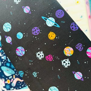 May include: Black decorative paper with a colorful space-themed pattern. The design features planets in shades of blue, purple, yellow, and pink, along with white stars. The paper is likely used for crafts or gift wrapping.