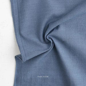 May include: A close-up of a blue fabric with a textured appearance. The fabric is folded and draped, creating soft shadows and highlights. The word "FABLEISM" is printed in white at the bottom of the fabric.