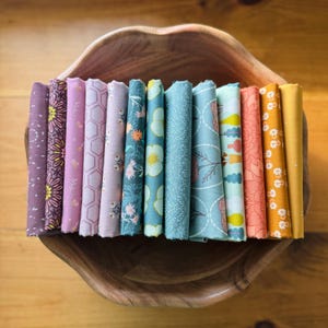 May include: A collection of folded fabric pieces in various colors and patterns, including floral, geometric, and abstract designs. The fabrics are arranged in a wooden bowl, showcasing a range of hues from purple and pink to teal and yellow. Ideal for quilting or crafting.