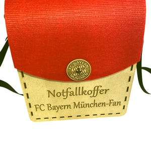 Könnte beinhalten: Eine braune Holzkiste mit einem roten Filzdeckel und einem schwarzen Riemen. Die Kiste hat ein goldenes Metallemblem mit dem Text "FC Bayern München" und einem Logo. Die Kiste hat den Text "Notfallkoffer FC Bayern München-Fan" auf der Vorderseite eingraviert.