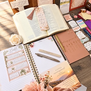May include: An open Bible with a floral bookmark, a spiral-bound Bible reading tracker, a notebook with the text God says you are, two pens, a cross ornament with text, a calendar, and various stickers featuring religious quotes.