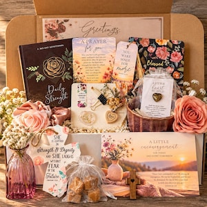 May include: A gift box containing a Daily Strength devotional book, a prayer card, a floral notebook, a heart-shaped stone in a pouch, a small pink glass vase with dried flowers, a wooden cross, a bag of treats, and cards with inspirational text.