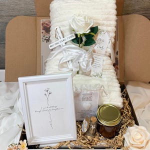 Puede incluir: Una caja de regalo que contiene una manta blanca texturizada, una rosa artificial blanca, un marco blanco con el texto I can do all things through Christ who strengthens me, un frasco pequeño con tapa dorada, un corazón y una cruz de madera.