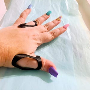 May include: A black thumb splint with a cutout design. The splint is worn on the left hand, which is resting on a light blue surface. The hand has painted fingernails in various colors.