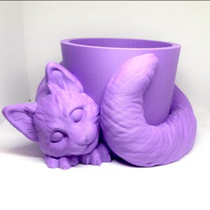 May include: A purple ceramic planter shaped like a cat curled up with its tail around a pot. The cat has closed eyes and a sweet expression.