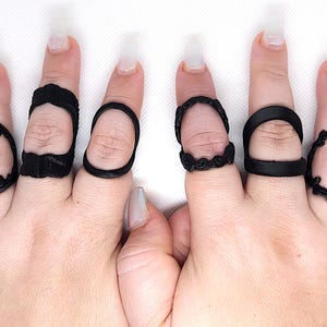 May include: A set of six black finger rings with different designs. The rings are made of a smooth, matte material and have a variety of shapes and sizes.