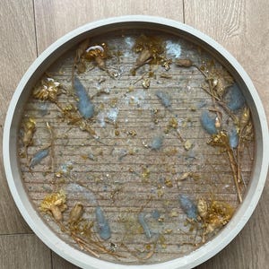 May include: A circular tray with a white border and a wooden base, featuring dried flowers, gold leaf, and pale blue elements. The tray is filled with clear resin, creating a decorative item. A decorative home decor item.