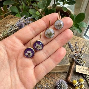 May include: Three round drop earrings featuring pressed white flowers encased in clear resin. Two earrings have deep purple backgrounds, while the third has a light lavender background. Each piece is set in a gold-toned metal frame with a hook.