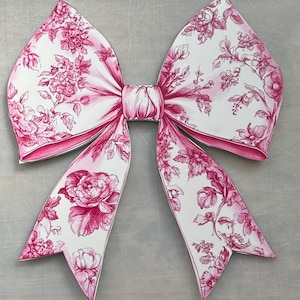 May include: A large, decorative bow with a floral pattern in shades of pink and white. The bow features a detailed design of flowers and leaves, with a solid pink border. The bow is a perfect accent for gifts or decorations.
