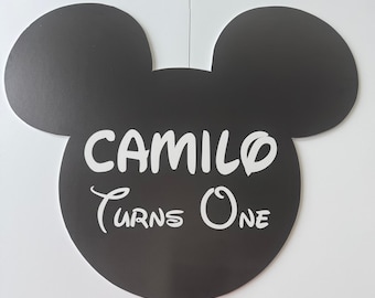 May include: A black Mickey Mouse head-shaped decoration with the text "CAMILO Turns One" in white. The design is a simple, bold graphic, perfect for a birthday celebration. The ears are large and rounded.