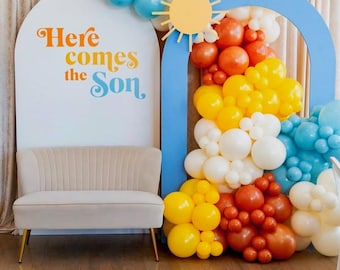 May include: A white arch with the text "Here comes the Son" in orange and blue lettering. A blue arch with a yellow sun cutout is decorated with a rainbow of balloons in blue, white, orange, and yellow.