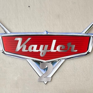 May include: A red and chrome emblem with the name "Kayler" in silver script. The emblem has a red background with silver trim and two silver fins extending upwards. The number "2" is at the bottom.