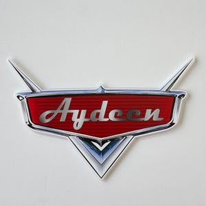 May include: A red and silver emblem with the name "Aydeen" in a stylized font. The emblem has a classic car design with a red background, silver accents, and a white border. The emblem has two silver wings.