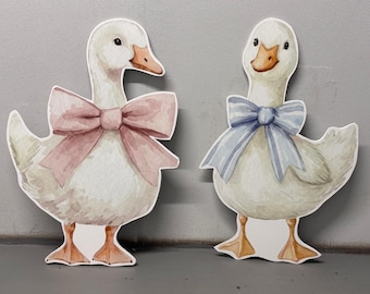 May include: Two decorative goose cutouts. One goose wears a pink bow, the other a blue bow. The geese are white with watercolor-style details, including orange beaks and feet. The background is a neutral gray.