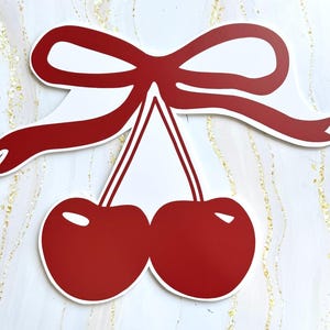 May include: A flat, die-cut decorative sign featuring two vibrant red cherries hanging from thin stems, topped with a large, stylized red ribbon bow. The design has a clean white outline and a smooth, solid red color finish on both the fruit and the bow.