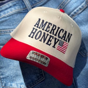 May include: A white and red baseball cap with the text "AMERICAN HONEY" embroidered in blue on the front. A small American flag is embroidered on the right side of the cap.