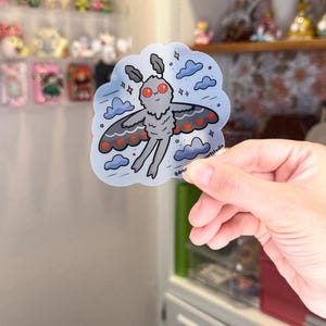 May include: A whimsical sticker featuring a cartoon moth with gray wings, red accents, and red eyes, floating among blue clouds. The sticker has a translucent background and the text "@dreamartstudio".