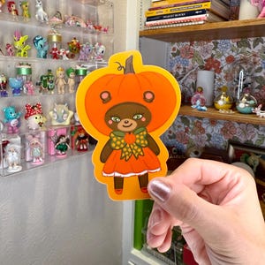 May include: A die-cut sticker featuring a bear character dressed in a pumpkin costume. The sticker has an orange pumpkin frame, a brown bear face, and an orange dress. The bear has a green bow and red shoes. The sticker is held in front of a shelf with figurines.