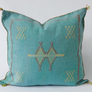 May include: A square throw pillow in a vibrant turquoise blue fabric featuring a woven texture. The center displays a geometric diamond pattern in yellow and red, with four smaller matching geometric motifs in the corners and small tassels.
