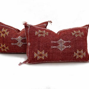 May include: Two rectangular throw pillows in a deep brick red colour, featuring intricate geometric embroidery in white and gold thread. Each pillow has small tassels at the four corners and a textured fabric surface with a repeating pattern.