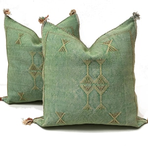 May include: Two square sage green throw pillows featuring a central geometric embroidery pattern in a lighter olive thread. Each pillow has small tassels at the four corners and a textured fabric finish with visible stitching along the edges.