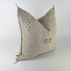 May include: A square throw pillow cover made of a textured, light beige fabric. The front features embroidered geometric patterns in white and tan thread. The edges have small fabric tassels at the corners and a visible metal zipper on the side.
