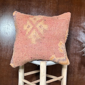 Puede incluir: Un cojín cuadrado y texturizado en tonos melocotón y naranja, con un patrón geométrico descolorido. El cojín está colocado sobre un pequeño taburete de madera clara. El fondo es una superficie oscura y texturizada.