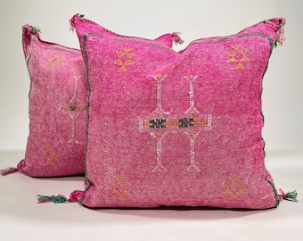May include: Two square throw pillows in a vibrant fuchsia pink color. Each pillow features a textured fabric surface with embroidered geometric patterns in yellow, white, and green. The edges are finished with dark green piping and small tassels.