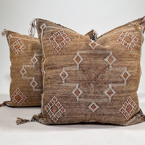 May include: Two square brown throw cushions featuring a textured fabric surface with white embroidered geometric diamond patterns and small brown accents. Each corner of the cushions is finished with a small decorative tassel detail.