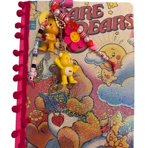 Może przedstawiać: Notatnik spiralny z okładką Care Bears z holograficznym brokatem, ozdobiony różową lamówką z pomponami. Metalowe kółko utrzymuje kolekcję zawieszek, w tym dwie figurki żółtych misiów, różowy kwiatek oraz różnorodne kolorowe koraliki.