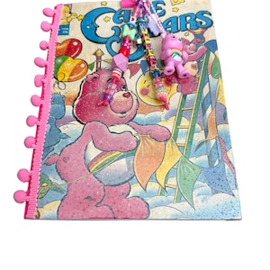 May include: A Care Bears notebook featuring a pink pom-pom trim on the left edge and a colorful beaded charm attached to the top. The cover displays a pink bear holding a rainbow banner, with a small pink bear figurine hanging from the charm.