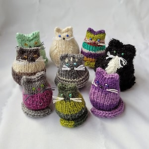 Peut inclure: Neuf jouets en forme de chat tricotés dans différentes couleurs et motifs, notamment vert, violet, gris, blanc et noir. Chaque chat a des traits du visage brodés et des yeux en perles.