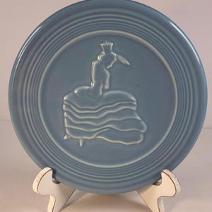 May include: A light blue ceramic plate with a raised relief design of a woman in a dress. The plate has concentric rings around the edge and is displayed on a white stand.