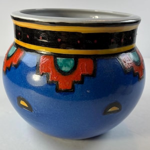 May include: A blue ceramic pot with a wide opening and rounded body. The pot features a black, yellow, and red geometric design around the rim and red and green patterns on the body. The pot has a glossy finish.