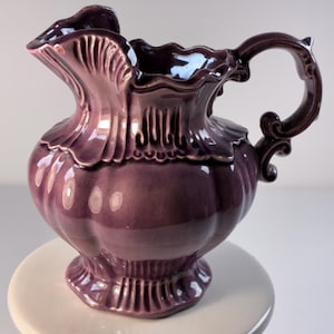 May include: A decorative, purple ceramic pitcher with a ruffled rim and ornate handle. The pitcher has a bulbous body and sits on a small, round base. The pitcher is approximately 8 inches tall.