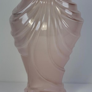 May include: A tall, elegant vase in a soft pink hue. The glass vase features a unique design with flowing, curved lines that create a draped effect. The vase has a wide base and a narrower neck, adding to its graceful appearance.