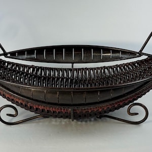 May include: A decorative, oval-shaped fruit basket with a dark brown woven design. The basket has a metal frame with decorative scrollwork handles and feet. The interior has a reddish-brown accent.
