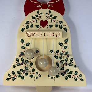 May include: A bell-shaped wooden wall hanging with a cream-colored base, featuring a red bow at the top, green holly leaves, red berries, and a central red heart. The word GREETINGS is printed in red, with three white cords holding wooden spheres.