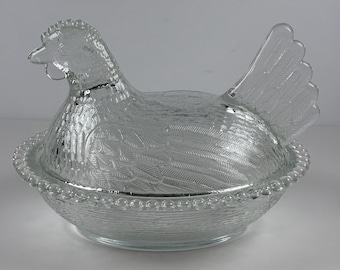 May include: A clear pressed glass hen on a nest dish. The lid is shaped like a hen with detailed feather textures, while the base features a beaded rim and a woven basket pattern. The entire piece is made of transparent, colorless glass.