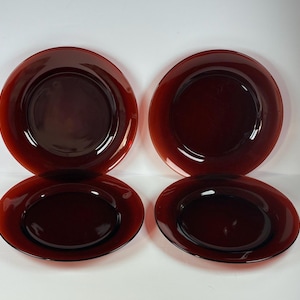 May include: Four circular dinner plates made of translucent dark red glass. Each plate features a smooth, reflective surface with a wide rim and a slightly recessed center, showcasing a uniform deep burgundy color throughout the glass material.