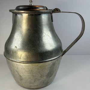 May include: A vintage metal pitcher with a hinged lid and a curved handle. The pitcher has a bulbous body and a silver finish. The lid is topped with a small decorative finial. The pitcher is likely made of pewter or a similar metal.