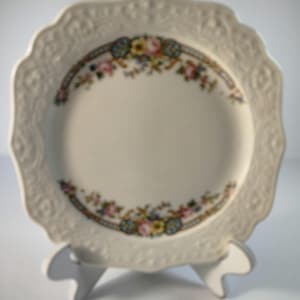 May include: A white ceramic plate with a scalloped edge and a floral design around the rim. The plate is on a white stand.