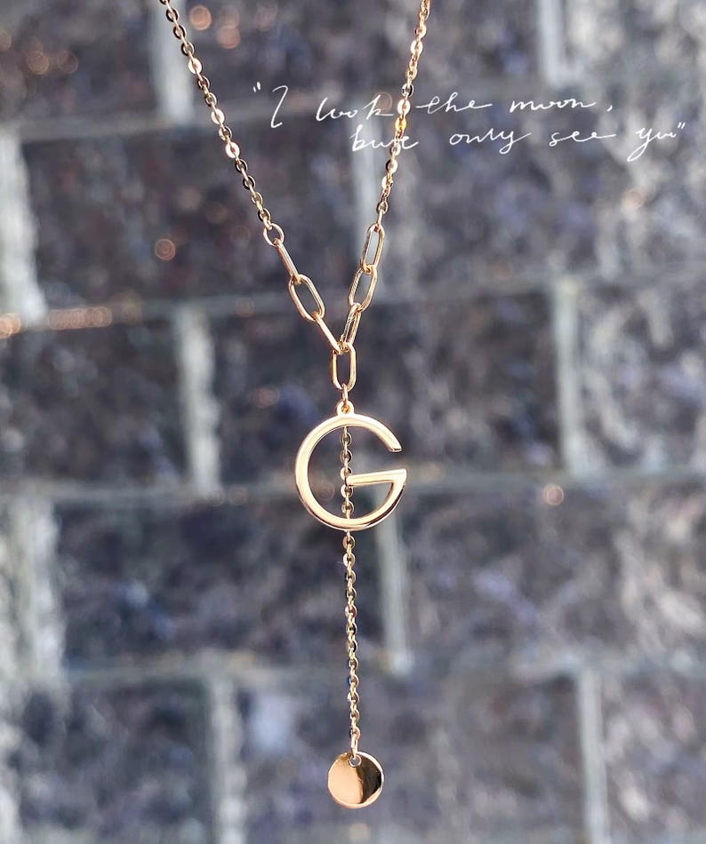 Puede incluir: Collar dorado con un colgante en forma de la letra "G". La cadena es fina y tiene un peque&ntilde;o dije circular. El texto "I look the moon, but only see you" es visible en el fondo.