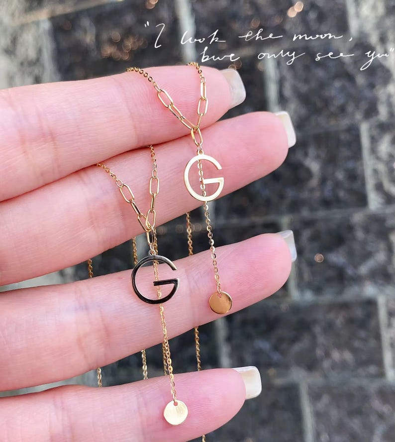 Puede incluir: Dos collares dorados con colgantes con la letra "G". Uno de los colgantes est&aacute; delineado en negro. Cada collar tiene una cadena delicada y un peque&ntilde;o dije redondo. La imagen incluye el texto: "I look the moon, but only see you."