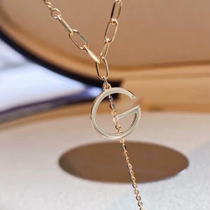 Puede incluir: Collar dorado con un gran colgante con la letra "G". El collar tiene una cadena delicada con un peque&ntilde;o dije circular. El dise&ntilde;o incluye diferentes estilos de cadena, creando un aspecto en capas. La joya se muestra sobre un fondo suave.